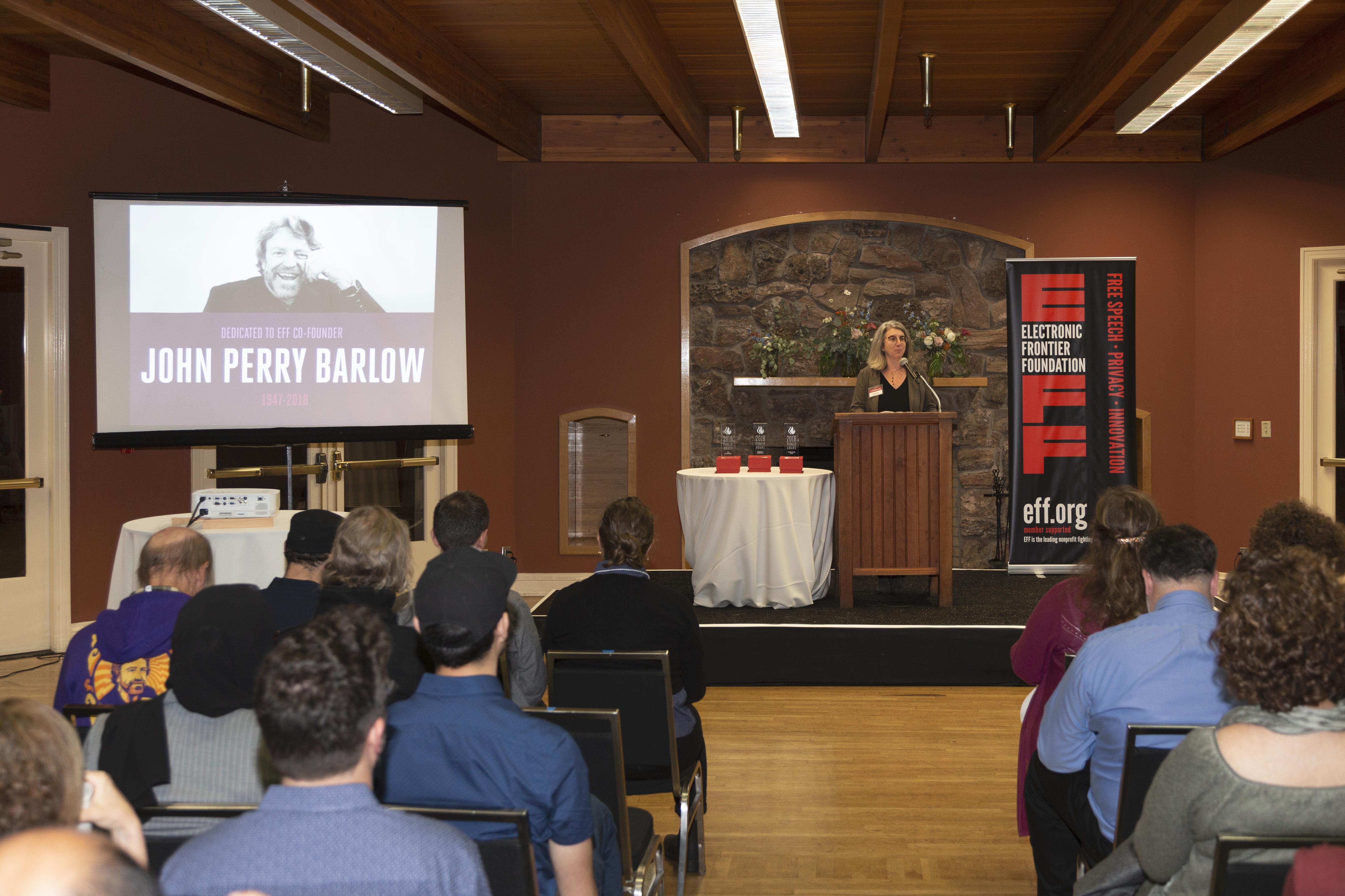 Cindy Cohn dedicates the Pioneer Award to EFF co-founder John Perry Barlow. Picture of screen and raised dias with podium. To the left, is a projected image of our co-founder, John Perry Barlow. At the poduim on the right is our Executive Director Cindy Cohn, opening the "Barlow" dedication.