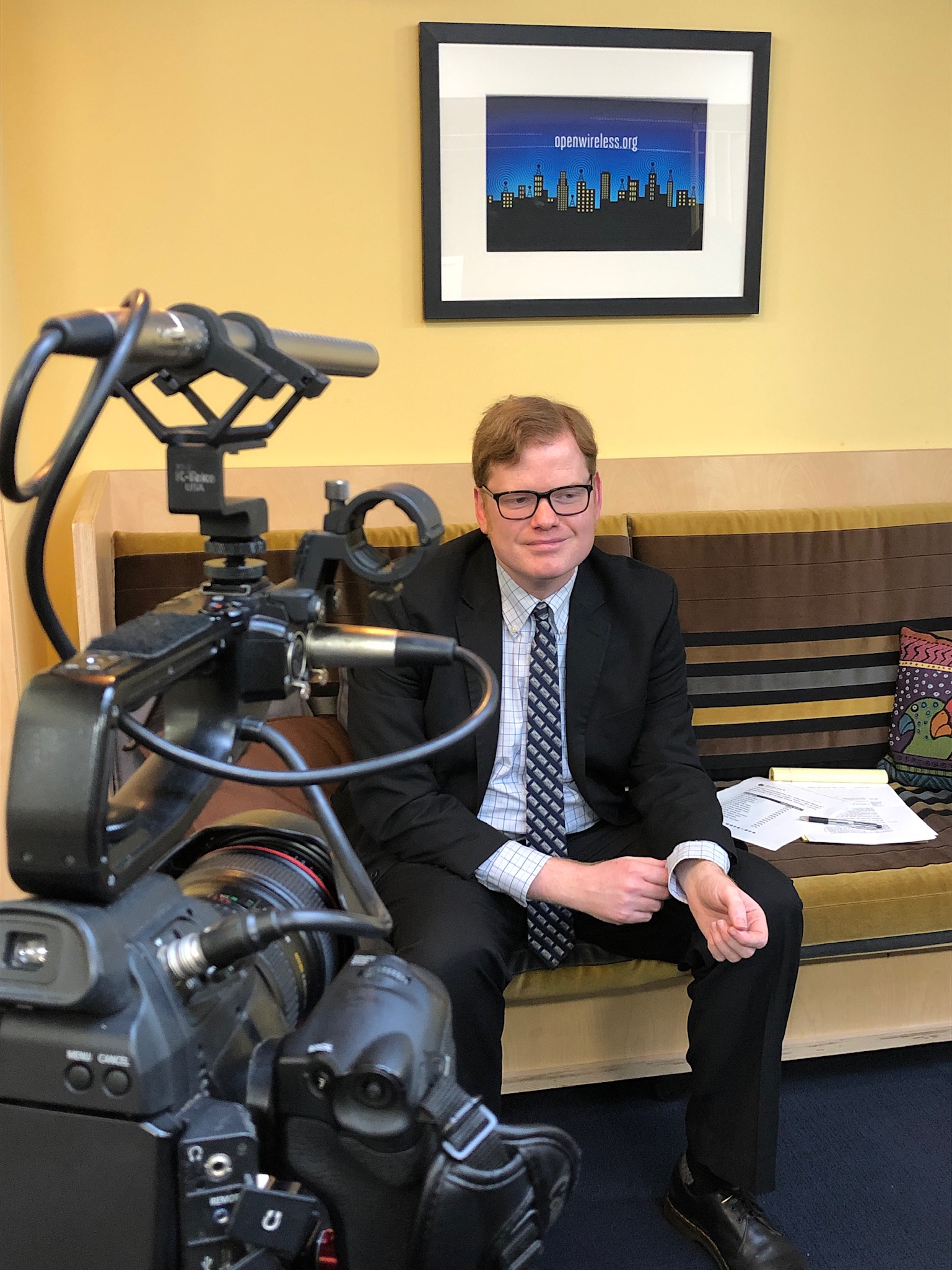 A photo of Elliot in a suit at the EFF office, in front of a television camera. Elliot is wearing his glasses, a suit and a blue tie underneath an illustration for Open Wireless.